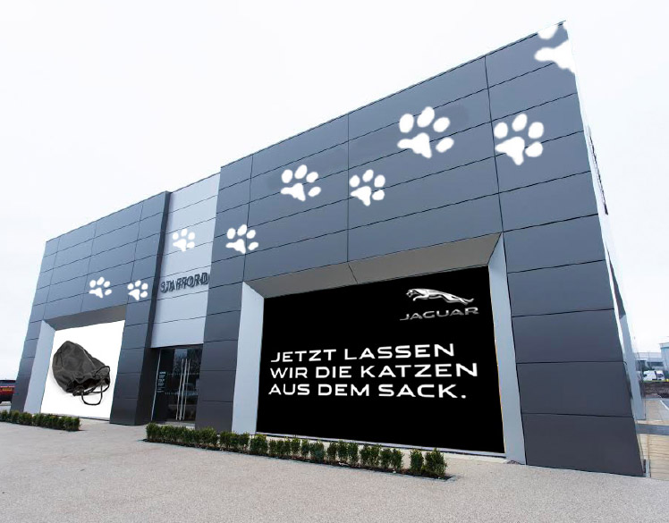 An image of a newly built car dealership. 
                One large window is covered with a sign showing an empty black bag. The other window has the headline Jezt 
                Lassen Wir Die Katzen Aus Dem Sack. There are large white paw prints moving up across the face of the building.
