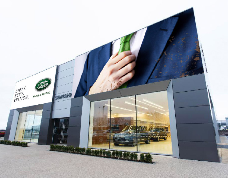 An image of a newly built car dealership. 
                A large sign above one window shows the headline Dirty. Sexy. British. A large sign above the 
                other window shows a close up of a person wearing a mud-splattered blue suit with a green tie.