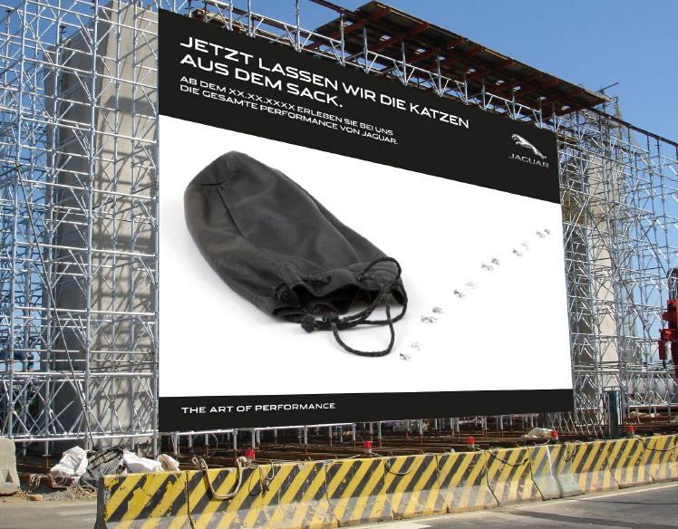 An image of scaffolding at a construction site. 
                The scaffolding is covered with a sign showing an empty black bag with paw prints leading out of it and 
                the headline Jezt Lassen Wir Die Katzen Aus Dem Sack.
